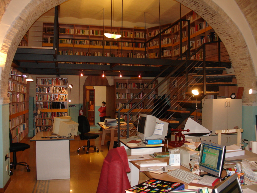 Sala interna della biblioteca comunale con scaffali, scrivanie ed una scala che porta ad un soppalco con altri scaffali di libri